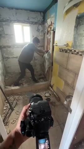 A person is seen in the background, wearing a dark t-shirt and dark pants, and is actively engaged in demolition work in a bathroom. They are using a cloth to clean debris from what appears to be a shower area. The bathroom itself is in a state of disrepair, with plaster and tiles removed from the walls, exposing the underlying brickwork and construction materials. Debris is scattered across the floor, suggesting an ongoing renovation project. In the foreground, a camera is held, pointed towards the person and the bathroom, implying that the scene is being filmed or documented. The lighting suggests it is daytime, with natural light entering from a window. The overall environment depicts a construction site within a bathroom.