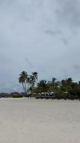 The video shows a wide, empty, white sand beach with palm trees and resort buildings in the background. The sky is overcast and gray. The camera pans slowly from right to left, capturing the expanse of the beach, the tropical vegetation, and the distant structures. There are no people or animals visible in the frame. The overall atmosphere is tranquil and serene, despite the cloudy weather.