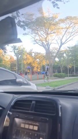 The scene is captured from inside a car looking out at a park on a sunny day. The car's dashboard is visible with a radio display showing some text. Outside, there's a park with a playground featuring swings, a slide, and other play structures. People are scattered around the playground, some walking and others sitting. A man on a green bicycle rides past the car, in front of the playground. Trees with full green foliage line the park providing shade. The overall setting seems relaxed and leisurely.