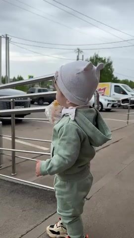 A baby stands outside in front of a building, eating from a pastry wrapped in plastic. The baby is wearing a green sweat suit with matching athletic shoes and a light grey knit cap. The baby’s face has crumbs on it. The building is next to a street with several parked cars. The sky is overcast. The baby looks at the camera then turns to look to the side. The building has a grey concrete facade with a silver railing.
