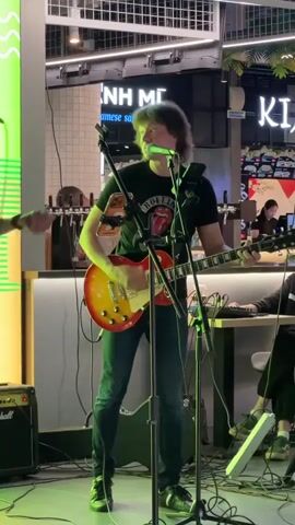 This video captures a live musical performance by a duo. A male guitarist, wearing a black t-shirt with a Rolling Stones logo and dark jeans, stands center stage, playing a sunburst-finished electric guitar and singing into a microphone. His long, brown hair frames his face as he engages with the music. To his left, a female singer with blonde hair, wearing a leopard-print dress, holds a microphone and smiles, seemingly in rhythm with the music.

The scene is set indoors, likely at a venue with a stage or performance area. Behind the performers, a counter with beer taps is visible, suggesting a bar or restaurant setting. The background also features signs with Vietnamese text, including "NH MI" and "japanese sandwich," indicating a potential Vietnamese food establishment. The lighting appears to be artificial, with vibrant green and yellow hues illuminating the space. The overall atmosphere seems lively and engaging, with both performers exuding enthusiasm. The presence of audio equipment like microphones and amplifiers confirms the musical event. The time of day is not explicitly indicated, but the indoor setting and artificial lighting suggest it could be evening or a dimly lit daytime venue.