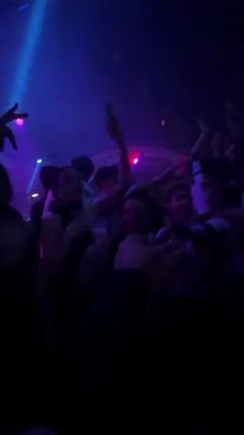 The video shows a group of young adults partying in a nightclub. The scene is dimly lit with colored lights, including blue and purple beams. Some individuals are on the dance floor, moving and gesturing to the music, while others are holding drinks and engaging with each other. One person is elevated above the crowd, making a hand gesture. Some are holding up cellphones, possibly recording the event. A bottle of Moet and Chandon champagne can be seen. A fog or smoke effect is used throughout the video.