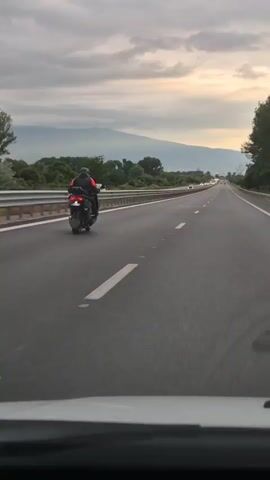 The video shows a view from inside a vehicle, looking forward. A motorcycle is in the right lane of a highway, ahead of the vehicle. The motorcycle rider is wearing a red jacket and a helmet. The highway has multiple lanes and guardrails on the left. In the distance, there are mountains and a cloudy sky with some light. Trees and vegetation are visible on the sides of the highway. The video appears to be taken at dusk.