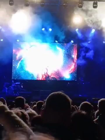 A view from behind a crowd of people at a concert. On the stage is a large screen displaying what appears to be a nebula or star cluster. A live band plays in front of the screen. There are blue spotlights and smoke illuminating the stage. The crowd is focused on the performance.