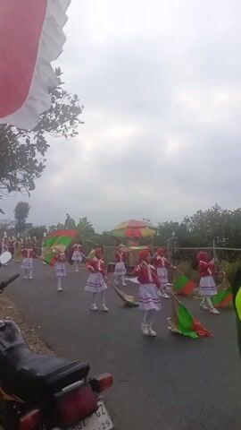 A group of young people in matching red and white uniforms, some wearing headscarves, are marching in a parade on a paved road. They are holding and waving flags that are green and red. Some individuals are also holding what appear to be poles. In the background, there are trees, a fence, and a colorful umbrella-like structure. A motorcycle is parked in the foreground. The sky is overcast.