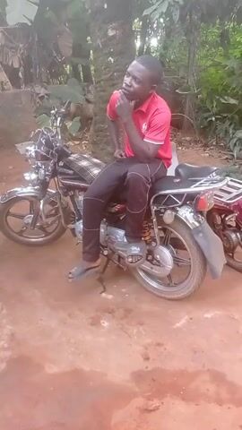 A man is sitting on a motorcycle, looking off to the left. The motorcycle is chrome colored with a plaid seat. The man is wearing a red shirt and dark pants. He is resting his chin on his hand and appears to be deep in thought. Behind the motorcycle is another motorcycle, maroon in color. The ground is red dirt. The scene appears to be outdoors, with green foliage in the background.