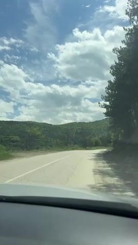 The camera is mounted inside a vehicle driving on a paved road through a rural area.  The road ascends slightly into a wooded area. The sky is visible above the trees and is a bright blue with fluffy white clouds.  The road is cracked.  A pile of dirt and rocks sits on the right shoulder of the road.  A portion of the dashboard is visible at the bottom of the frame.  A brief, abrupt exclamation of surprise is audible at around 0:03.