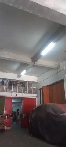 The video shows an interior space with a concrete ceiling and exposed beams. Fluorescent lights are mounted on the ceiling. A row of windows with metal grates is visible above a red sliding door. There are objects hanging behind the grates. A dark object, possibly covered with a tarp, is in the foreground on the right. The environment appears to be a storage area or a workshop.