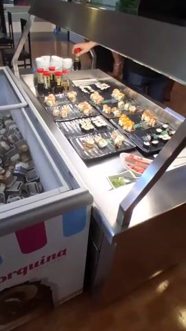 The video pans across a buffet line in a brightly lit establishment, likely a restaurant or cafeteria. The scene showcases a variety of food items arranged in stainless steel trays and containers.

In the foreground, to the left, a large glass-fronted freezer is filled with individual portions of what appears to be ice cream or frozen desserts. The branding on the freezer, "la Menorquina," is visible. Adjacent to the freezer, a counter displays an assortment of sushi rolls arranged on black platters, with small bottles of soy sauce and other condiments readily available. A hand is seen reaching for one of the soy sauce bottles.

Further down the buffet line, the camera reveals numerous containers filled with colorful fruits, including sliced watermelon, pineapple, and mixed fruit salads. Other sections offer what seem to be small cups of yogurt or pudding, some with individual packaging. There are also trays with savory items like small pastries or fried snacks, and a tray of brownies. Plates are neatly stacked in shelves below the counter.

The overall atmosphere suggests a self-service dining setting, possibly during a mealtime. The lighting is artificial and bright, indicating an indoor environment. The presence of various food stations, from sushi to desserts and fruits, suggests a diverse offering. No specific time of day can be determined, and there are no weather cues. The visible text "la Menorquina" on the freezer indicates a potential brand of ice cream being served. The general mood conveyed is one of abundance and a well-stocked buffet.