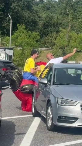 Two men stand near a silver car in a parking lot. The man on the left wears a yellow shirt, blue pants, and a black cap. He is partially inside a large, inflatable red, blue, and yellow costume. The man on the right wears a white t-shirt and is holding a phone up, appearing to take a photo or video. Another person is visible through the car's passenger side window. A black car is parked to the left of the silver car. In the background, there are trees and what appears to be an electrical charging station.