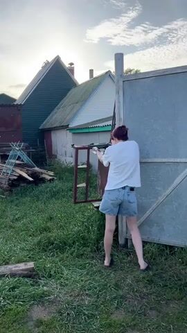 A woman stands in a grassy backyard, wearing a white t-shirt and denim shorts. She holds a black rifle and shoots it.  A man approaches her, and she gives him the middle finger.  In the background, there are wooden buildings, a ladder, and a metal fence.  The sky is cloudy with patches of blue.