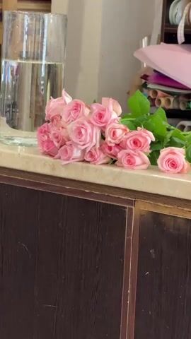 In this video, a woman is arranging a bouquet of pink roses. She is in a brightly lit room with a counter and shelves stocked with floral supplies, including rolls of decorative paper and ribbons. The counter is made of a light-colored material with a subtle pattern, and a dark wood cabinet is visible below it. A large glass vase filled with water sits to the left of the roses. A small yellow object, possibly a tool, is also on the counter. The woman's focus is on the roses, suggesting she is a florist or preparing flowers for an event. The scene appears to be indoors, likely in a flower shop or a crafting area. The lighting is even and bright, without any strong shadows, indicating it could be daytime or under artificial light. There is no visible text in the media file.