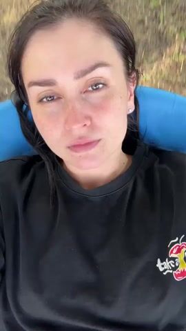 This is a close-up, overhead shot of a woman lying on her back. She has dark hair pulled back, is wearing a black t-shirt with a graphic on the lower right side, and has freckles. She is lying on a blue surface, which appears to be some kind of inflatable object. The background is blurred and consists of green and brown grass and leaves, suggesting an outdoor setting. The woman is looking directly at the camera with a neutral expression. The video is short, and the camera appears to be stationary. There are no other people or significant objects visible in the frame.