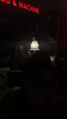 The dimly lit video captures a recording session. A shirtless man stands by a drum set under a small lamp, while another individual in a baseball cap plays an acoustic guitar in the foreground. A red neon sign reading "OO & MACHINE" hangs above. Red curtains are visible to the right.