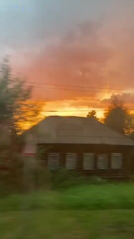 A low-angle, handheld video captures a blurry, sunset landscape from a moving vehicle. The sky is filled with vibrant orange and pink clouds, suggesting the sun is setting. In the foreground, a grassy field is visible, with trees and a rustic wooden house. The motion blur suggests the camera is moving horizontally, possibly out a window, as the scenery shifts from left to right. A power pole or similar vertical structure is also present in the frame. The overall impression is one of a serene, rural sunset.
