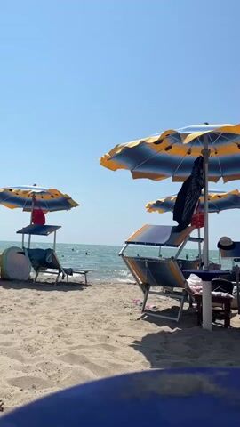 The video opens on a beach with sand in the foreground and the ocean in the background. Several lounge chairs and umbrellas are set up on the sand. In the distance, the ocean is visible with a few people in the water. The sky is clear and blue. The camera pans slightly to the right, revealing more of the beach and the ocean. There are no specific actions or interactions captured in this brief clip, other than the natural movement of the waves. The overall impression is a peaceful, sunny day at the beach.
