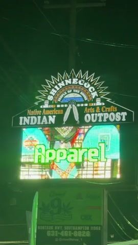 The video shows a brightly lit sign at night. The sign has multiple levels. The top part of the sign reads "SHINNECOCK" in large letters with "PEOPLE OF THE TRIBE" written below it. Below that, it says "Native American Arts & Crafts" and "INDIAN OUTPOST". The bottom part of the sign is a digital display showing various images, including the word "Apparel" in large, green, pixelated letters. Below the digital display is a smaller green sign with white lettering that reads "Cloud 9 Sauce" and includes a phone number and website address. The overall scene is illuminated by the bright lights of the sign, set against a dark night sky. The lighting is predominantly green.