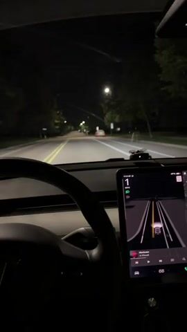 The video shows a first-person view from inside a Tesla driving at night on a suburban road in Vernon Hills, United States of America. The driver's side steering wheel is visible in the foreground, and a large touchscreen display mounted on the dashboard shows the car's navigation system. The screen displays a futuristic visualization of the road ahead, with lane markings and other vehicles detected by the car's sensors. The current speed is displayed as 53 mph, and the time is indicated as 68 degrees Fahrenheit and 67% battery. Music is also playing, with the title "Hurricane" by "I Prevail" visible.

The scene is dark, with streetlights illuminating the road and the surrounding trees. The road appears to be a two-lane street with houses and trees lining either side. Several other vehicles are visible ahead, including a white car and a few cars with their brake lights on. The overall impression is one of a calm, autonomous drive through a quiet neighborhood at night. No people are clearly visible, and the focus is on the car's technology and the driving experience. The weather appears clear, and the mood is serene and perhaps a little futuristic given the technology on display. The location is identified as Vernon Hills, USA.