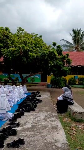 A wide-angle shot reveals a group of children sitting on a blue tarp in an outdoor setting, all dressed in white uniforms with head coverings. Rows of black shoes neatly line the edge of the tarp. A woman wearing a headscarf and black dress sits on a concrete ledge nearby, holding a baby wrapped in a patterned blanket. The backdrop features lush green trees and a school building with colorful artwork on its exterior. The sky overhead is overcast, suggesting a cloudy day. The camera gradually pans across the scene, capturing the entirety of the assembled group and the surrounding environment.