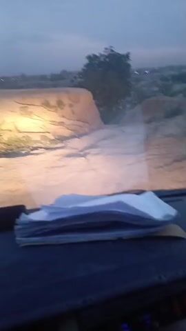 The video shows a view from inside a vehicle at night, looking out through the windshield. Headlights illuminate a sandy, uneven ground in front of the vehicle. Patches of green vegetation are visible on the ground. In the distance, under a dark blue sky, are faint lights that appear to be from a distant town or buildings. There's also a car dashboard visible in the foreground, along with a tissue box. A conversation can be heard in an unidentified language.