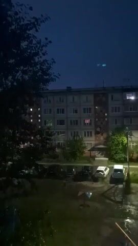 This is a vertical video taken at night of a residential building and its surroundings. The sky is dark blue, suggesting it is either dusk or pre-dawn. The building is several stories high and appears to be made of concrete. Numerous windows are visible, and some have lights on inside. In front of the building, there is a courtyard with several parked cars, including a white SUV and a dark-colored sedan. There are also some trees and bushes, and a small playground structure. The ground in the courtyard appears wet, suggesting it may have rained recently. The video is shaky, indicating it was likely filmed by hand.