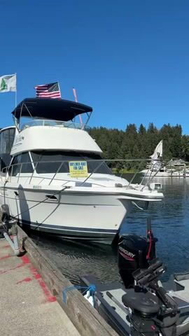The video shows a white Bayliner 4007 boat docked at a marina on a clear, sunny day. The boat has an American flag and another flag with a green tree on it flying from its mast. A "FOR SALE" sign is prominently displayed on the front of the boat. The camera pans to show the dock, which is made of wood and concrete, and other boats docked nearby. The water is calm and reflects the blue sky. The background features a line of trees and a bright blue sky.