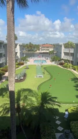 The video shows an eye-level view of the courtyard of a condominium complex, framed by a tall palm tree in the foreground. In the center of the courtyard, there is a putting green with red flags marking the holes. Next to the putting green is a shuffleboard court. Beyond the shuffleboard court is a swimming pool surrounded by lounge chairs and a white fence. Beyond the pool is a large house on the water. The courtyard is bordered on both sides by the condo buildings, which are painted white. The sky is blue with scattered white clouds. The lighting is bright, and the view is mostly static. There are no people visible in the video.