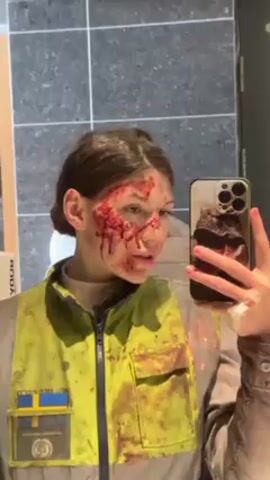 A young woman takes a selfie in front of a bathroom mirror. She wears a high-visibility yellow vest splattered with what appears to be blood. She has what looks like a large bloody wound around her right eye and cheek. She holds an iPhone with a brown phone case.  The bathroom has gray tile and a white box labeled “BODY” is visible on the counter. The woman looks directly at the camera and then slightly to her left.