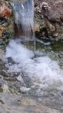 A close-up, low-angle shot shows water cascading from an unseen source into a shallow pool. The water is clear and appears to be flowing over rocks and dirt. The surrounding environment is rough and natural, with some greenery visible. The overall impression is one of natural beauty and tranquility.