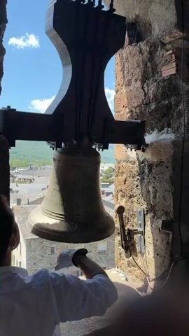 In a bell tower overlooking Benasque, Spain, a person wearing white gloves and a white shirt is operating a large bronze bell. The bell is suspended by a wooden beam and a dark metal mechanism, and it is swinging back and forth, creating a deep, resonant sound. The person is pushing the bell to set it in motion, with their gloved hands touching the sides of the bell. The scene is illuminated by bright sunlight, suggesting it is daytime. The sky is blue with scattered white clouds. Beyond the bell tower opening, a view of the town of Benasque is visible, with its stone buildings and tiled roofs, nestled amongst rolling green hills. The overall atmosphere appears to be peaceful and historic, highlighting the traditional practice of bell ringing in a picturesque European town. There is some faint text visible on the bell itself, but it is too blurry to be legible.