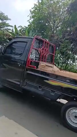 A black pickup truck is parked on the right side of the road, facing away from the camera. The truck is loaded with large cardboard boxes. The road is paved and appears to be an urban or suburban setting, with trees and vegetation visible in the background. A person on a motorcycle is seen riding on the road in the background, to the left of the pickup truck. The sky is visible and appears to be clear with some clouds. The lighting suggests it is daytime.