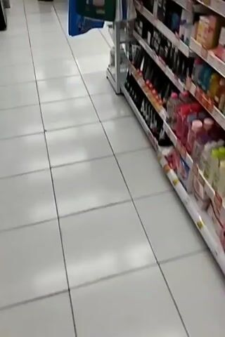 The video pans across a retail store. In the foreground, a green reusable shopping bag hangs from a display. To the right, shelves are stocked with various products. In the background, several people are visible. One person, wearing a blue shirt and dark pants, appears to be working behind a counter. Another person, wearing a white shirt and a headscarf, stands near the counter. Further back, another person in a white shirt and dark hat is also present. The floor is tiled and appears to be white. The lighting is bright, suggesting an indoor environment.