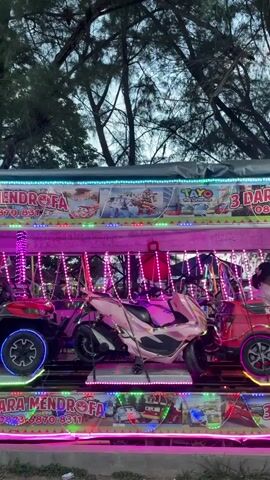 A low-angle shot shows a children’s amusement ride in operation. The ride consists of toy vehicles, including a red ATV, a pink motor scooter, and a grey car, moving along a track. Two young boys are seated in the vehicles, with one in the red ATV and another in the grey car. The ride is brightly lit with pink, blue, and green LED lights. Above the ride, a sign reads "3 DARA MENDROFA" with phone numbers. Trees are visible in the background. The sky appears overcast.