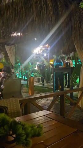 The video captures an outdoor or semi-outdoor restaurant or bar area at night, under a thatched roof. In the foreground, a wooden table and rattan chairs are visible, with two women seated at a table on the left. One woman wears a leopard print top. Balloons, including yellow and silver star-shaped ones, are visible in the background on the left. The midground features lush green plants illuminated by green lights, with a pathway or deck made of wooden planks. On the right side of the midground, several individuals are gathered around a bar or counter. One person, wearing a black shirt with white lettering (possibly "VORTEX"), stands behind the counter. Other people are standing in small groups, with some appearing to dance or sway to music. Bright artificial lights create lens flares, illuminating the scene from above.