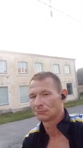 A man is looking at the camera. He has short, light brown hair and is wearing a black and yellow jacket with white writing on it. He also has a black earbud in his right ear. Behind him is a light-colored, two-story building with several windows. To the right of the building, there is a road and a tree. The sky is overcast. The man's expression is neutral. The video is filmed outdoors during the day.