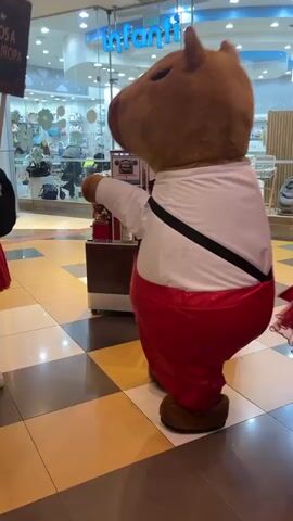 The video shows three anthropomorphic capybara mascots walking in what appears to be a shopping mall. Two are dressed in red dresses and one is dressed in red pants and a white shirt. The mascots are walking towards the right of the frame. The environment includes checkered tile flooring, store fronts, and people walking by.