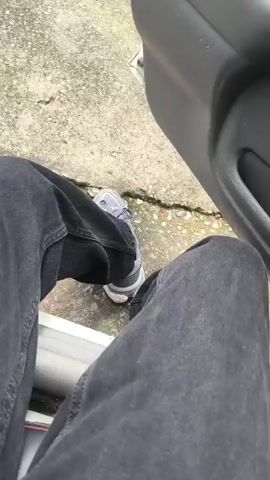 The video begins with a close-up shot of a person's legs, wearing dark pants and grey sneakers, next to an open car door. The camera then pans upwards and to the right, revealing the interior of a car and the exterior through the open door and window. Two cars are parked on a gravelly surface in the background, with trees and a blue sky with white clouds above. The view through the car window shows a hilly landscape with trees, buildings, and what appear to be laundry hanging on a line. The scene is outdoors, likely during the daytime, with good visibility.
