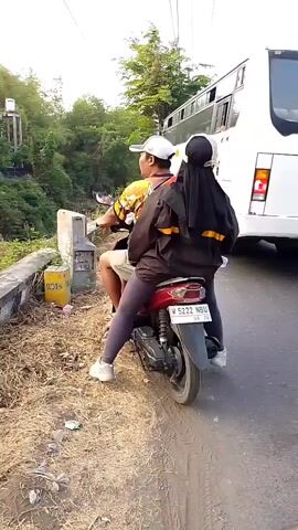 A motorcycle with two people is stopped on the side of a road, near a concrete barrier and overgrown vegetation. A large white bus is driving past the motorcycle on the road. The motorcycle is red and the people are wearing dark clothing. The person in the back is wearing a headscarf. A yellow sign is attached to the concrete barrier. The road appears to be wet. In the distance, more vehicles, including a large truck and other motorcycles, are visible on the road. The scene appears to be in a tropical or sub-tropical climate, with trees and lush greenery surrounding the road.