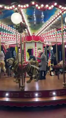 A merry-go-round is in motion. Riders, including children, sit on the animals as the carousel rotates. The ride is ornately decorated with lights and painted animals, including horses and a tiger. The backdrop includes a blue and white sky scene. People watch from the outside of the carousel. The lighting is bright and festive.