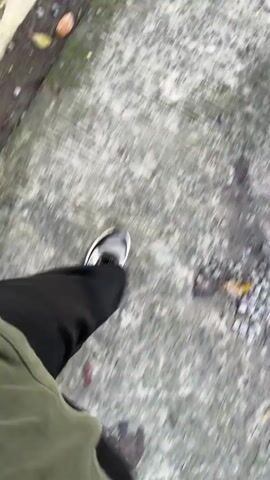 The video shows a person's legs and feet walking on a concrete path. The person is wearing black pants and black and white sneakers. The concrete path has cracks and is lined with some gravel and dry leaves. The footage is shot from a first-person perspective, looking down at the person's legs as they walk. The movement of the camera suggests the person is walking at a moderate pace. The environment appears to be outdoors, possibly a park or a trail.