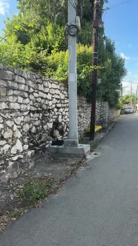 The video shows a street view on a sunny day. A stone wall runs along the left side of the road, topped with dense green foliage. A gray utility pole with wires stands in front of the wall, and a dark brown wooden pole is next to it. A bright yellow bench is visible against the stone wall, near the base of the wooden pole. The road itself is asphalt and appears to be an urban or suburban street. Further down the road, a white car is parked, and a few houses are visible on the right side. The sky is blue with some white clouds. The camera appears to be handheld and is moving slightly.