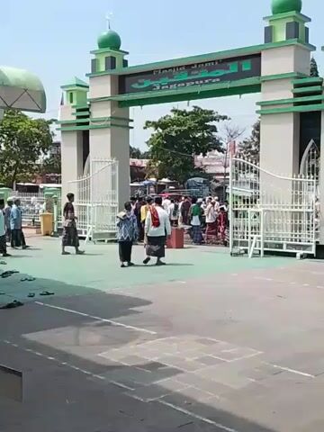 The video shows a wide shot of the entrance to Masjid Jami Jagapura, a mosque, under bright daylight. People are walking into and out of the gate of the mosque. The mosque's entrance is a large, decorated structure in grey and light green, topped with small green domes. Arabic text is written on the gate. The foreground shows a paved area with a few sandals scattered about. In the background, behind the gate, more people are visible, as well as vehicles and trees. The general atmosphere appears to be calm and orderly.