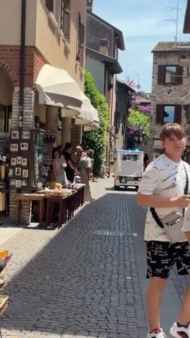 The video shows a narrow cobblestone street lined with buildings and shops. A white van with flashing blue lights is driving down the street. Several people are walking on the street, some are looking at the van. There is a display of goods on tables to the left of the street, and some people are looking at these goods. The buildings have balconies and windows, and there are some plants on the buildings. The sky is clear and the sun is shining.