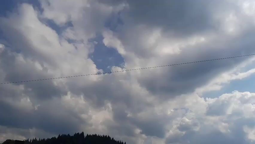 This is a still image taken from a video file. The scene depicts a wide shot of a cloudy sky. The clouds are a mix of white and gray, suggesting a dynamic weather pattern, possibly a partly cloudy day with potential for rain or just passing clouds. A thin, dark line, likely a power line or cable, stretches horizontally across the frame, creating a visual division of the sky. Below the sky, a dark, silhouetted treeline forms the bottom edge of the image, suggesting a forested or hilly area. The trees appear to be evergreen, with a dense, natural growth. The lighting suggests it is daytime, with the sun likely behind some of the clouds, creating varied illumination. The overall impression of the scene is natural and perhaps a bit dramatic due to the cloud formations. There is no visible text in the image. The location context provided is Gliczarów Górny, Poland, which, combined with the treeline, suggests a rural or mountainous landscape characteristic of that region. No people or specific objects beyond the power line are discernible. The activity, if any, is simply the natural movement of clouds in the sky.
