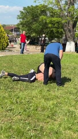 On a sunny day, two shirtless young men in dark pants wrestle on a grassy lawn. An older man in a blue shirt watches them closely. The shirtless men grapple on the ground before rising to their feet, still wrestling. A small crowd of onlookers stands in the background, near trees and a building.