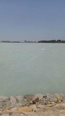 The video presents a view of a large body of water. The water has a milky, pale blue or greenish-gray color. Small waves and ripples are visible on the surface, indicating a slight wind or current. Along the right edge of the frame is a bank or shoreline consisting of rocky and sandy terrain. The rocks are various sizes and appear to be a mix of colors, including gray, brown, and tan. On the far shore, barely visible, are the faint outlines of what appear to be buildings. The sky above is a clear, pale blue. The image is oriented vertically.