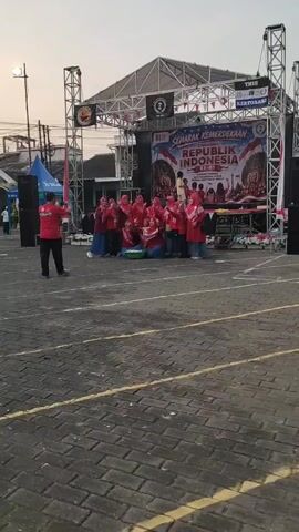 A group of people in red shirts and headscarves are posing for a photo in front of a stage. The stage has a banner that says "SEMARAK KEMERDEKAAN REPUBLIK INDONESIA KE-78". There are speakers and lighting equipment on either side of the stage. A man in a red shirt is standing in the foreground, facing the group and holding up a phone, possibly taking a picture. The ground is paved with interlocking bricks. The lighting suggests it is either early morning or late afternoon.
