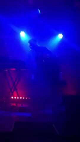 A musician is standing behind a keyboard on stage, backlit by blue stage lights. The musician is wearing what appears to be a light colored shirt and dark pants. There are other stage lights visible in front of the keyboard, creating a mix of blue and red lighting on the stage. The stage is otherwise dark.