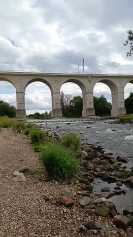 A wide shot captures a river flowing under a large, arched stone viaduct on a cloudy day. The river is shallow and rocky, with visible currents and rapids. Green grass and bushes line the riverbank on the left, adjacent to a gravel path. The viaduct has multiple arches, supporting what appears to be a railway track. Trees and vegetation are visible on the far bank of the river. The sky is overcast with dramatic clouds. The camera is stationary.