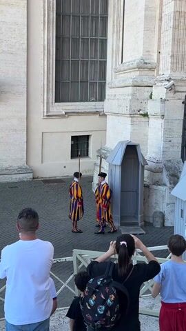 Two Swiss Guards stand on a stone-paved courtyard facing each other, holding spears. They are dressed in traditional striped uniforms of yellow, red, and blue. Behind them is a gray guardhouse and the side of a large stone building with windows. In front of the guards is a low white fence separating them from a small group of tourists. The tourists are looking at the guards, some taking pictures.  A man in a white shirt stands to the left, a woman with a backpack and her hair in a ponytail stands in the center, and two children are visible on the right side of the frame.